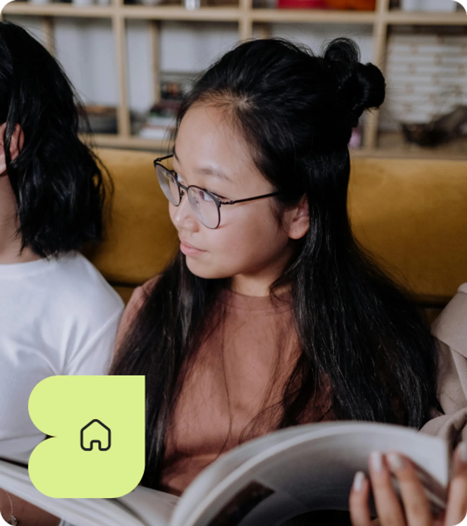 Young girl with long black hair and glasses reading a magazine while seated on a sofa.