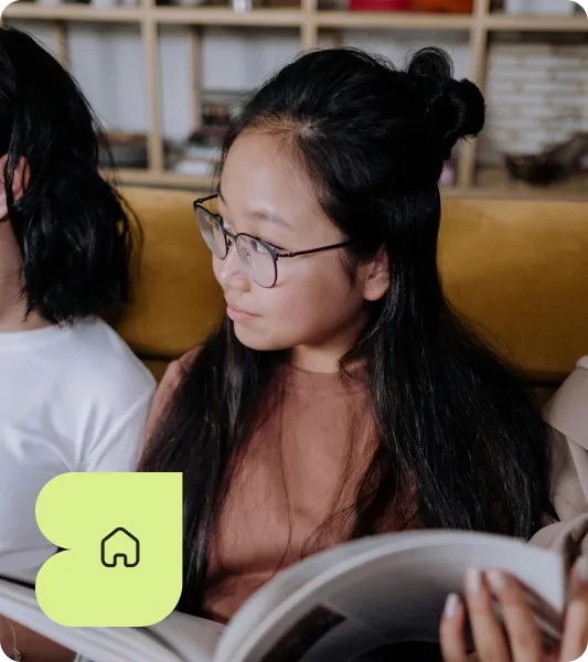 Young girl with long black hair and glasses reading a magazine while seated on a sofa.