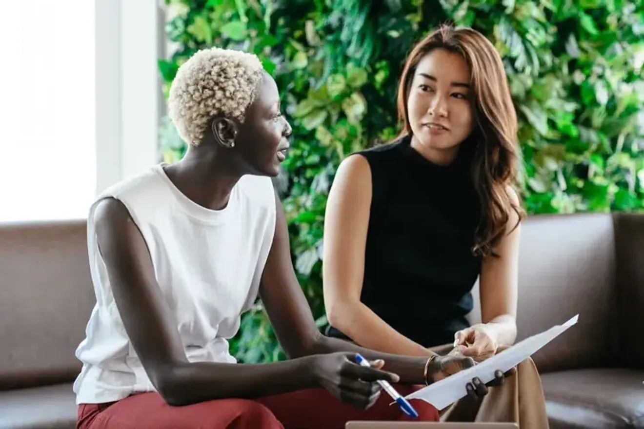 Two women having a business discussion, holding papers and pens, with a green plant wall in the background, representing collaboration and communication