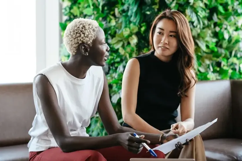 Two women having a business discussion, holding papers and pens, with a green plant wall in the background, representing collaboration and communication