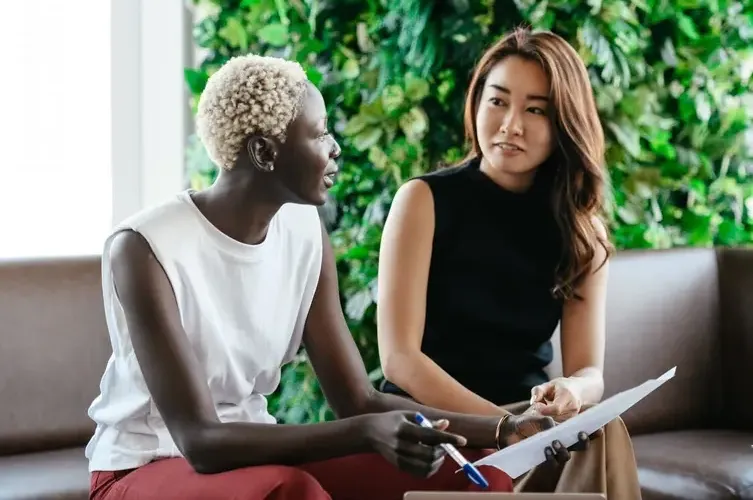 Two women having a business discussion, holding papers and pens, with a green plant wall in the background, representing collaboration and communication