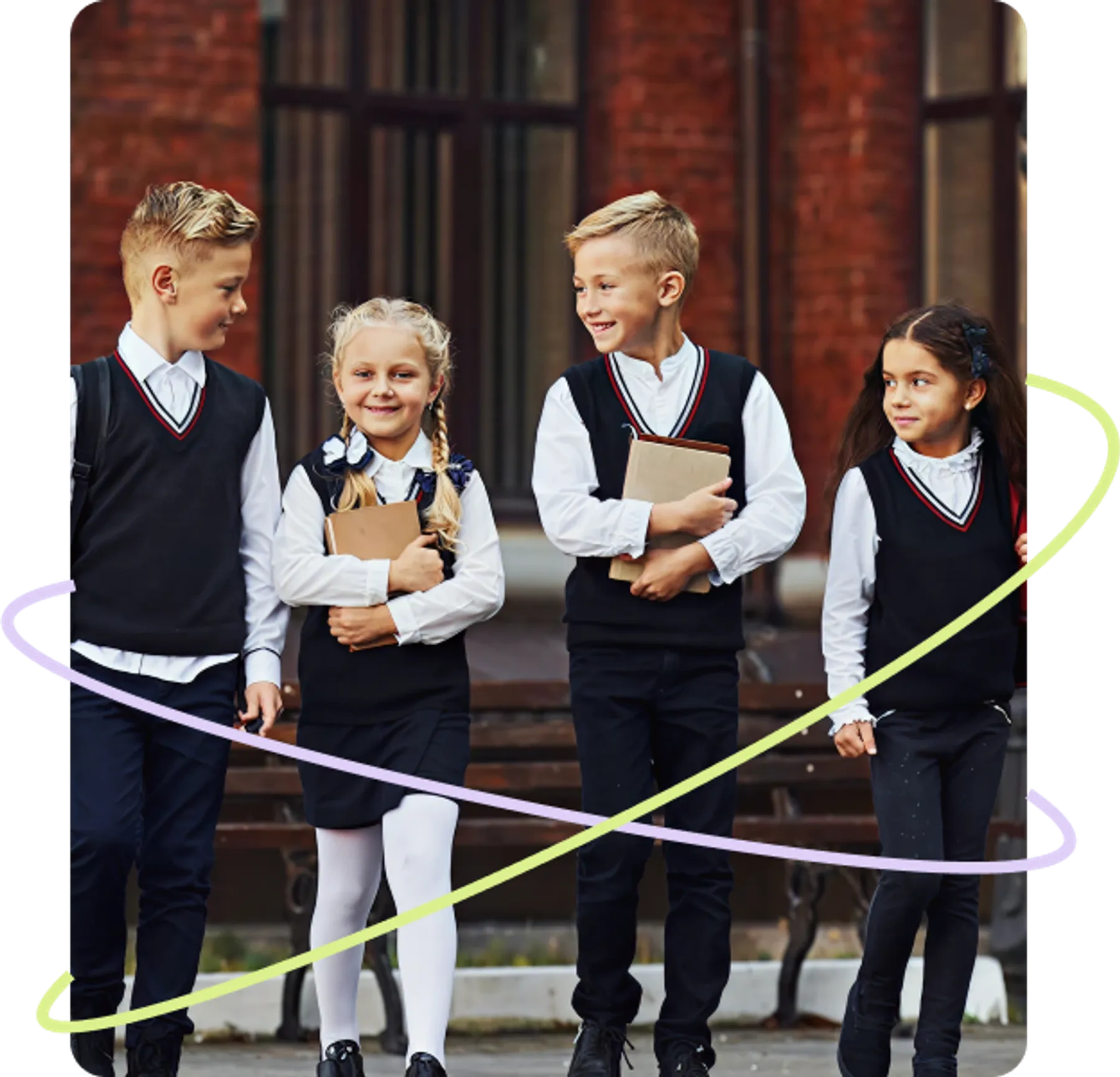 Four young school children in uniforms walking together outside a school building, carrying books and smiling.