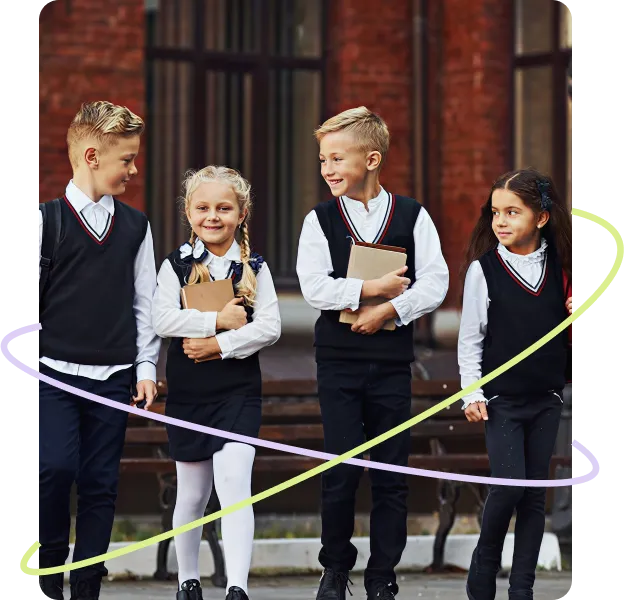 Four young school children in uniforms walking together outside a school building, carrying books and smiling.