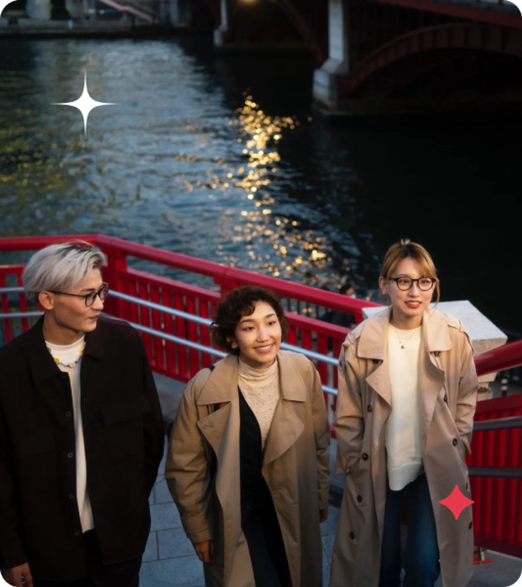Three casually dressed young adults walk and smile near a red railing along a riverfront at dusk.