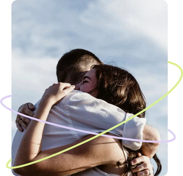 A woman with long dark hair hugs her partner tightly in an emotional reunion under a bright sky.