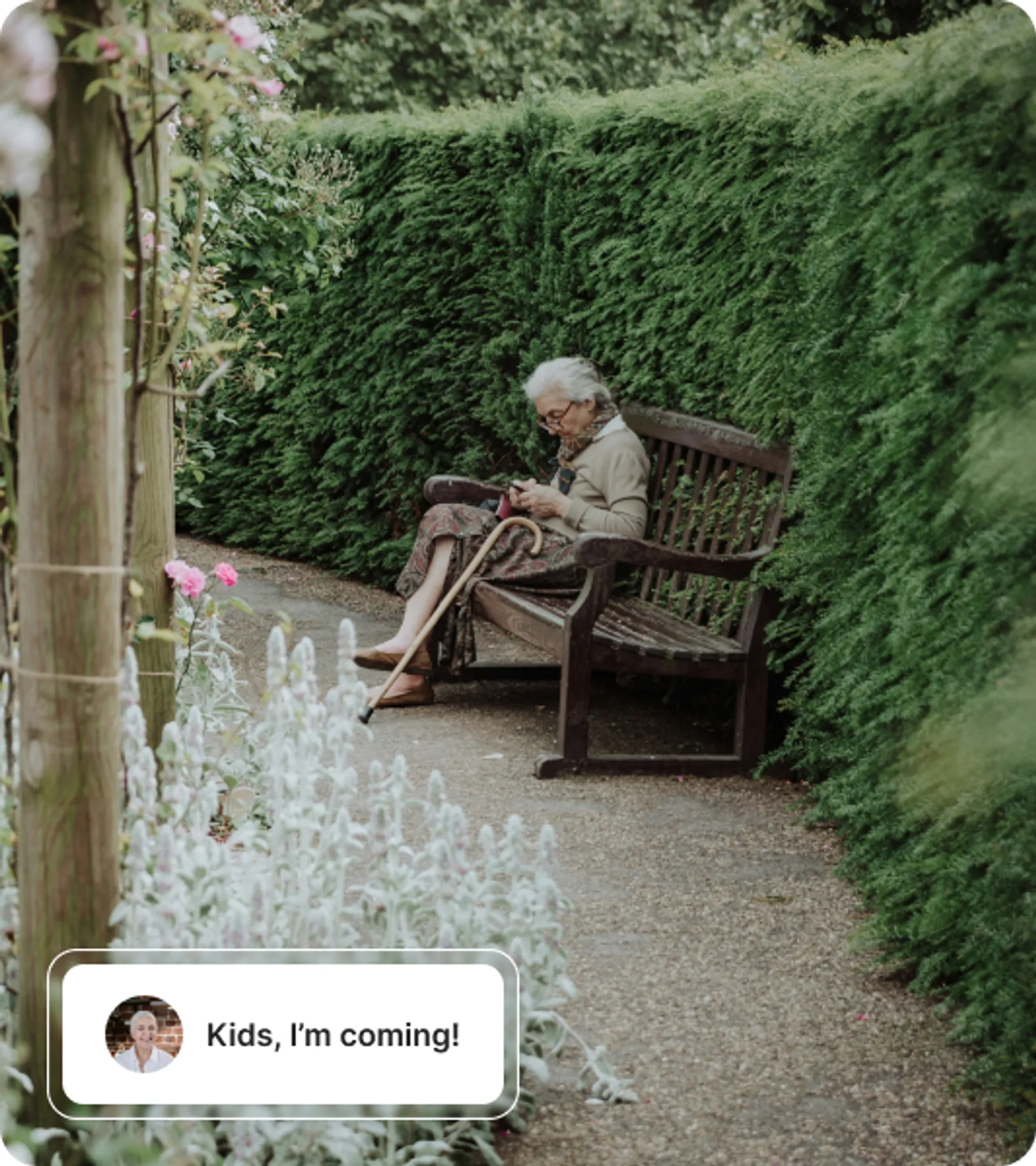 An elderly woman sits alone on a wooden bench in a peaceful garden path, using a mobile phone with a cane by her side.