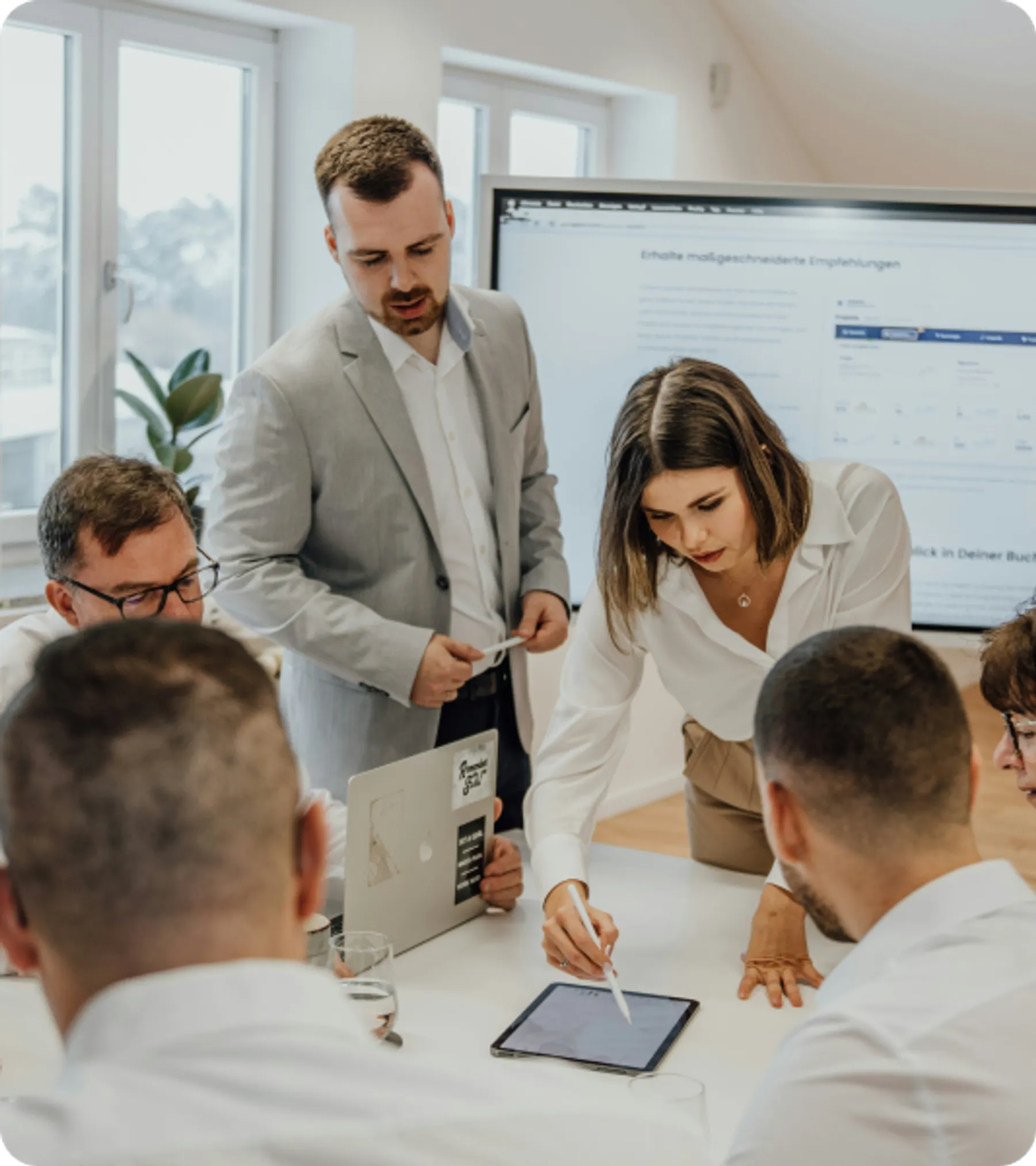 Group of professionals in a business meeting, with a woman leading a discussion and pointing at a digital tablet, symbolizing teamwork.