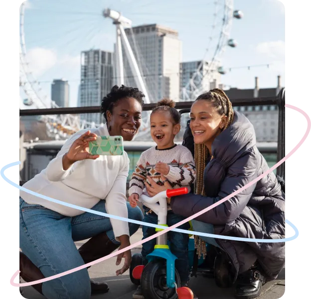 Two women and a smiling child take a selfie near the London Eye, with the child seated on a tricycle.