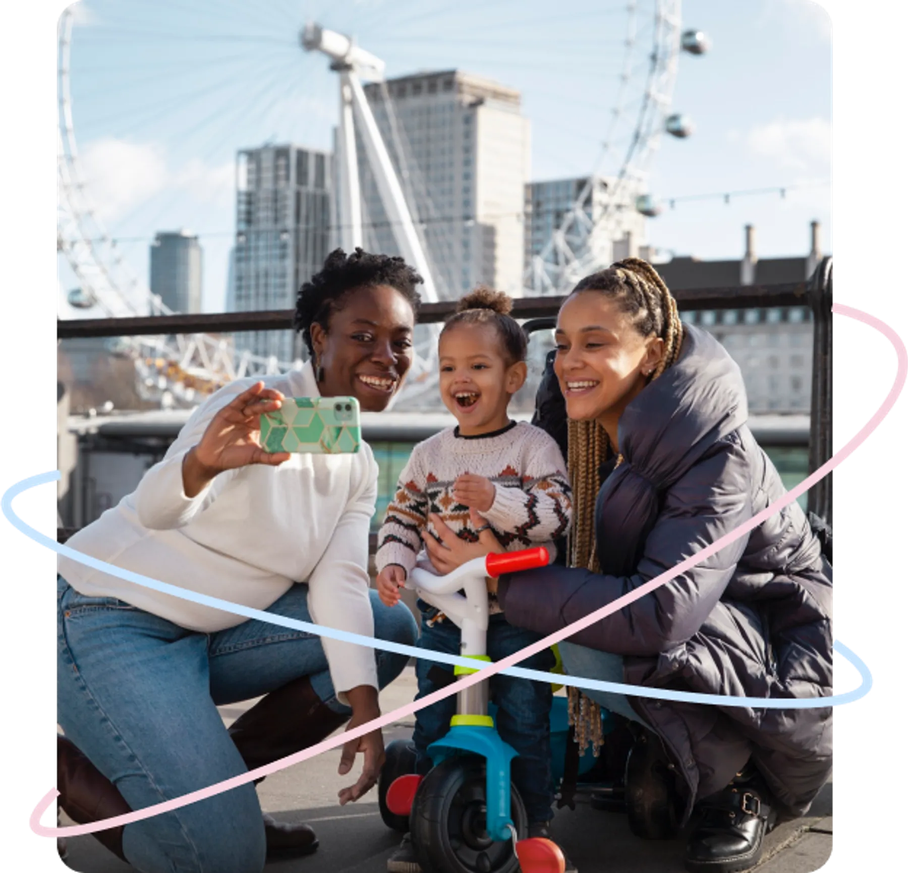Two women and a smiling child take a selfie near the London Eye, with the child seated on a tricycle.