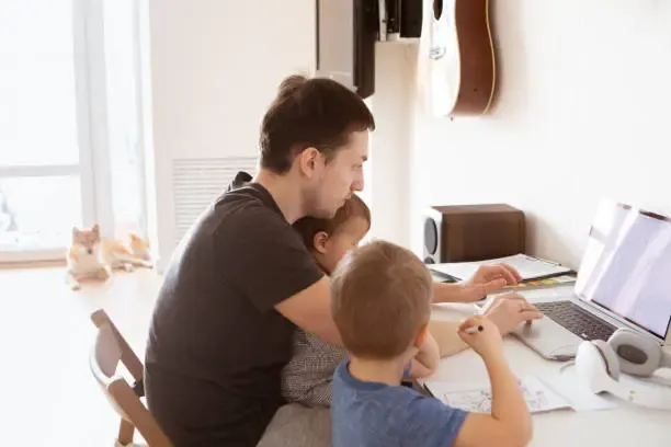 A father working on a laptop at home with two young children on his lap, balancing work and childcare