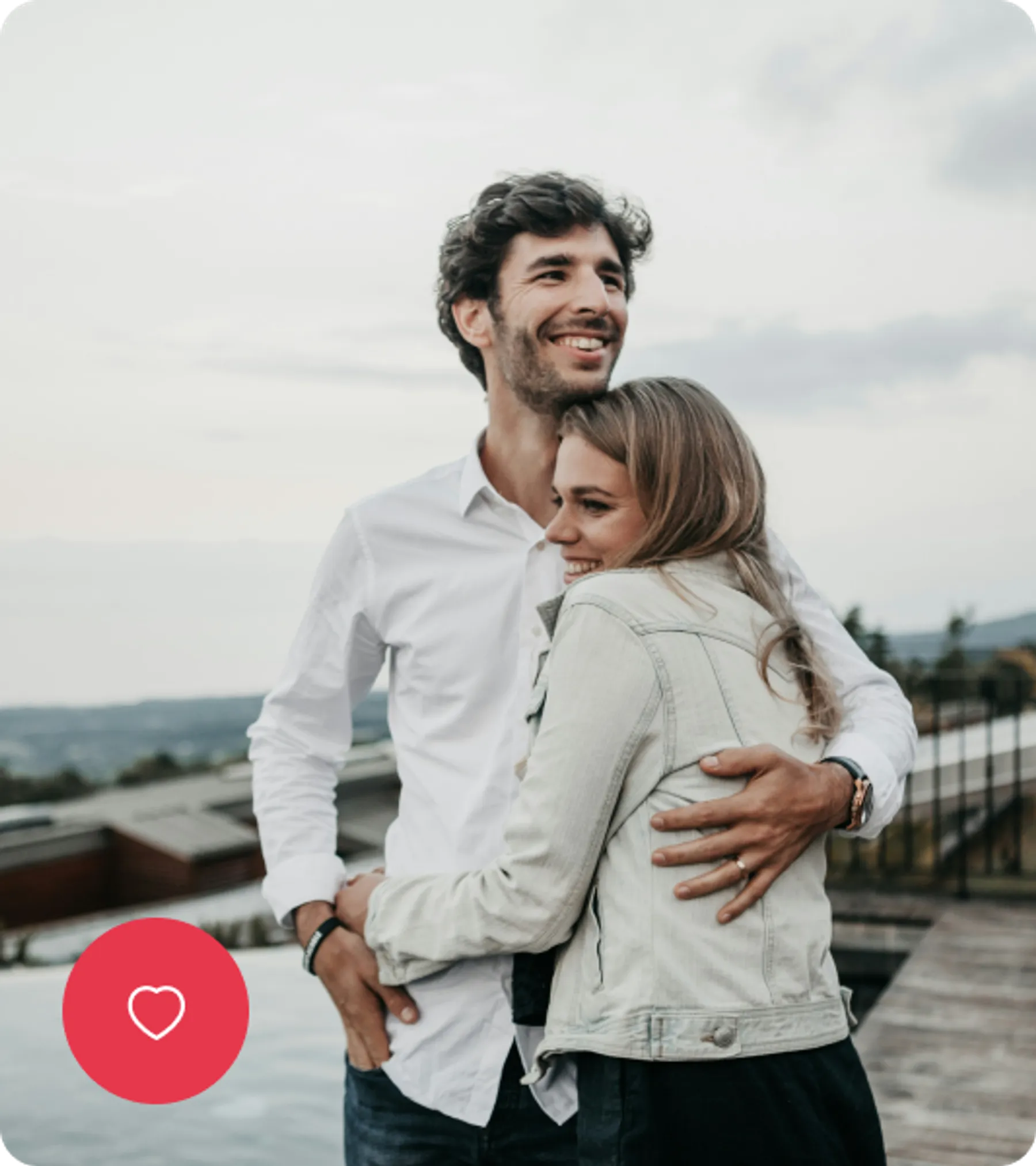 A smiling couple embraces outdoors on a cloudy day, standing near a scenic overlook with rooftops in the background.