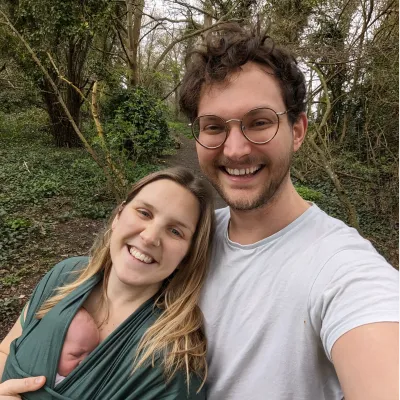 Smiling couple with baby in wrap carrier taking a selfie during a walk in the forest