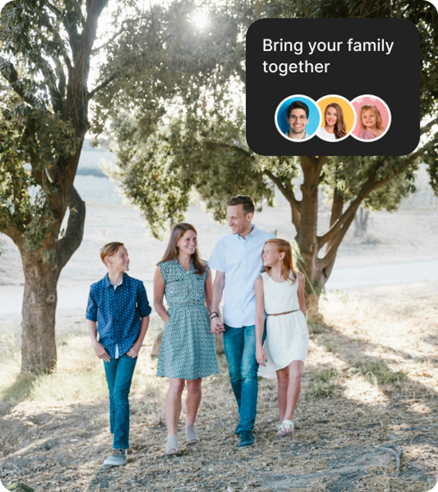 A smiling family of four with two parents and two children walking hand in hand along a sunlit path surrounded by trees.