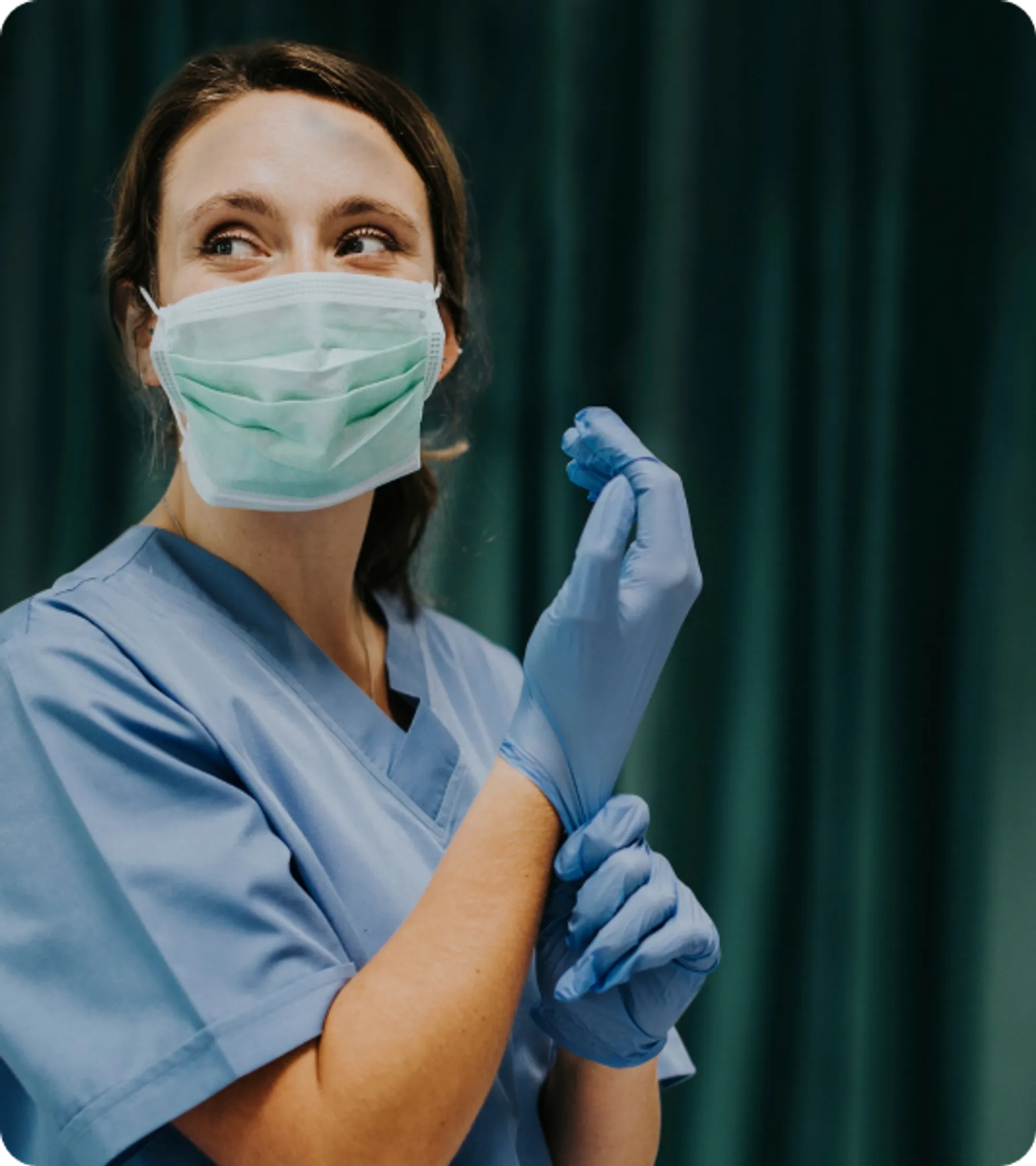 A female healthcare worker wearing scrubs, a surgical mask, and blue gloves, smiling while adjusting her glove in a clinical setting.