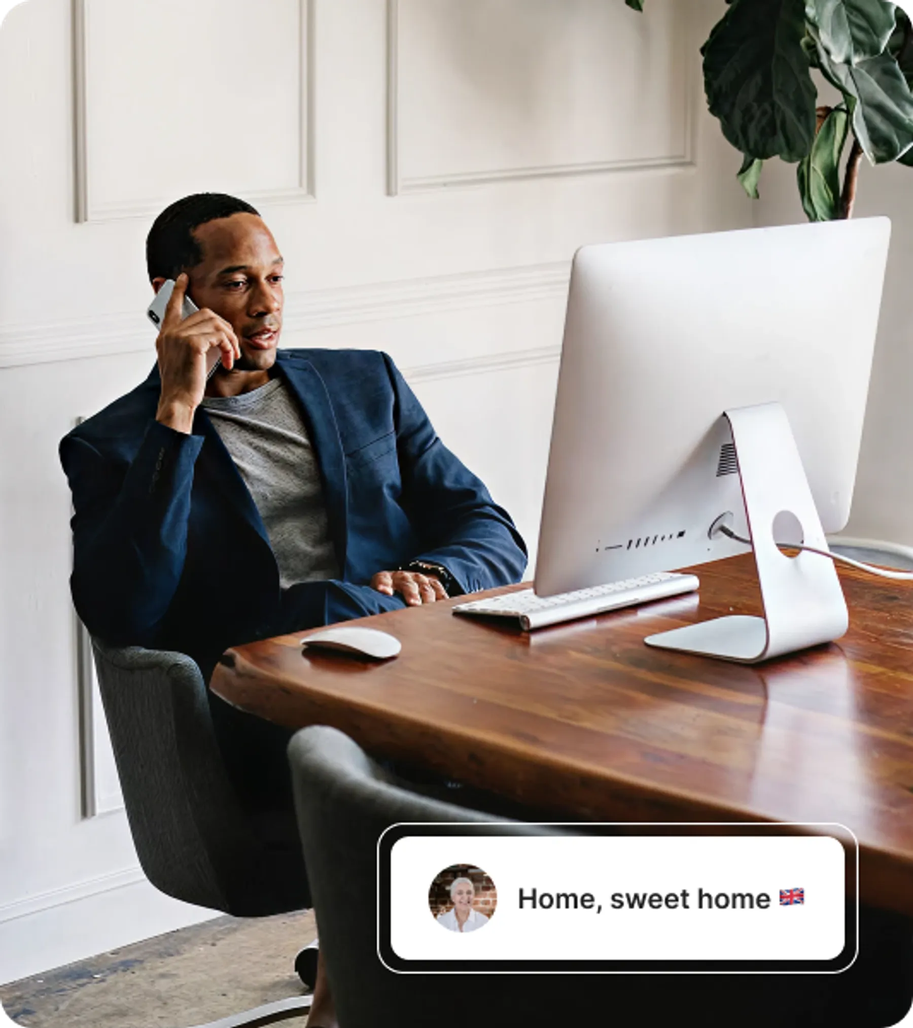Man in a blazer speaking on the phone while seated at a desk with a desktop computer in a home office.