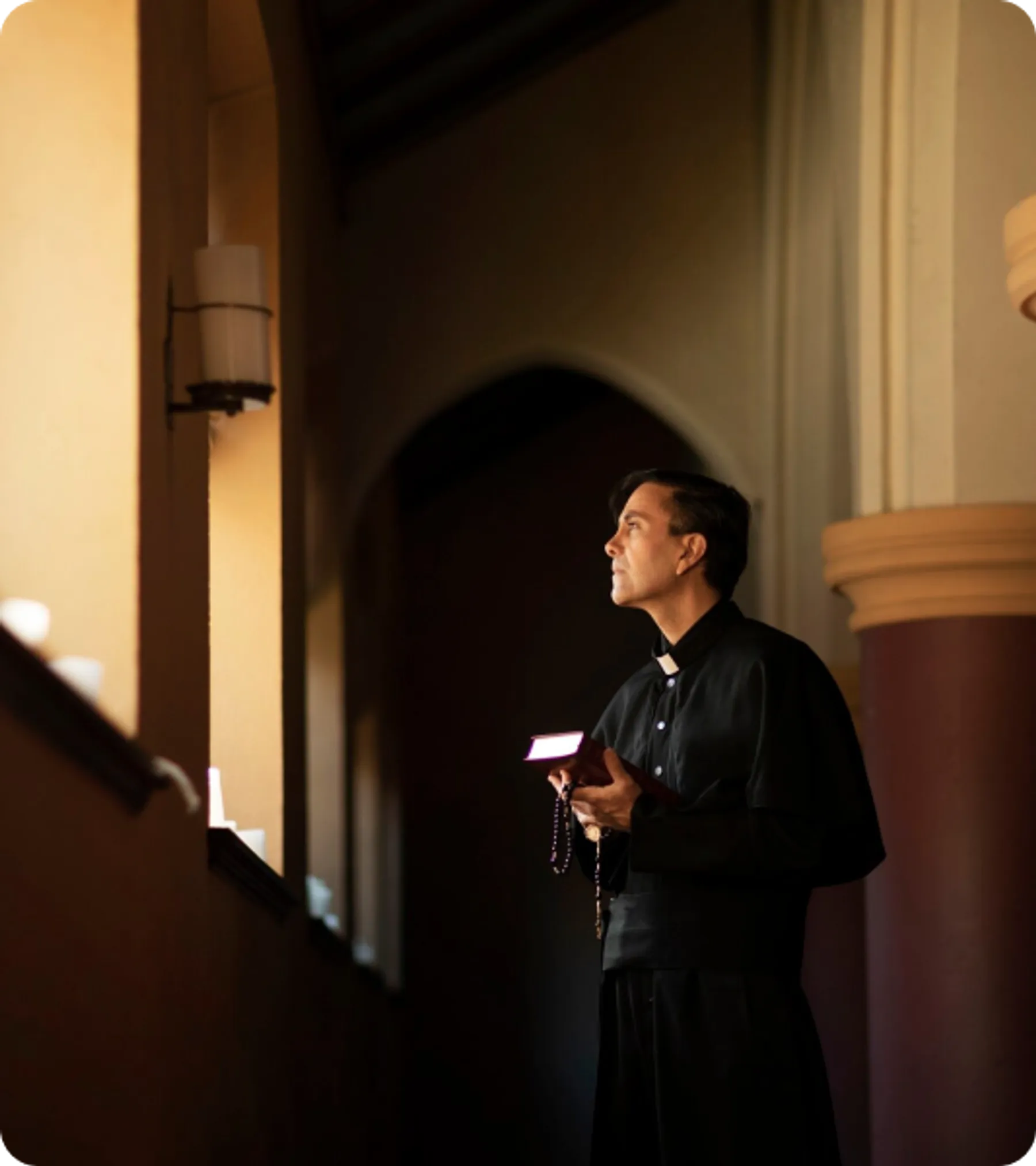 A male minister in black clerical attire holds a book and rosary while standing in prayer inside a softly lit church interior.