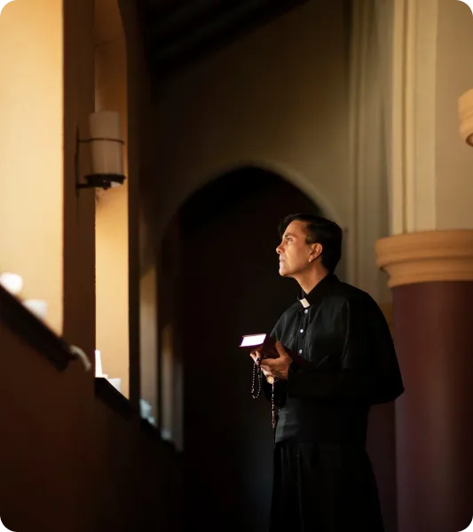 A male minister in black clerical attire holds a book and rosary while standing in prayer inside a softly lit church interior.