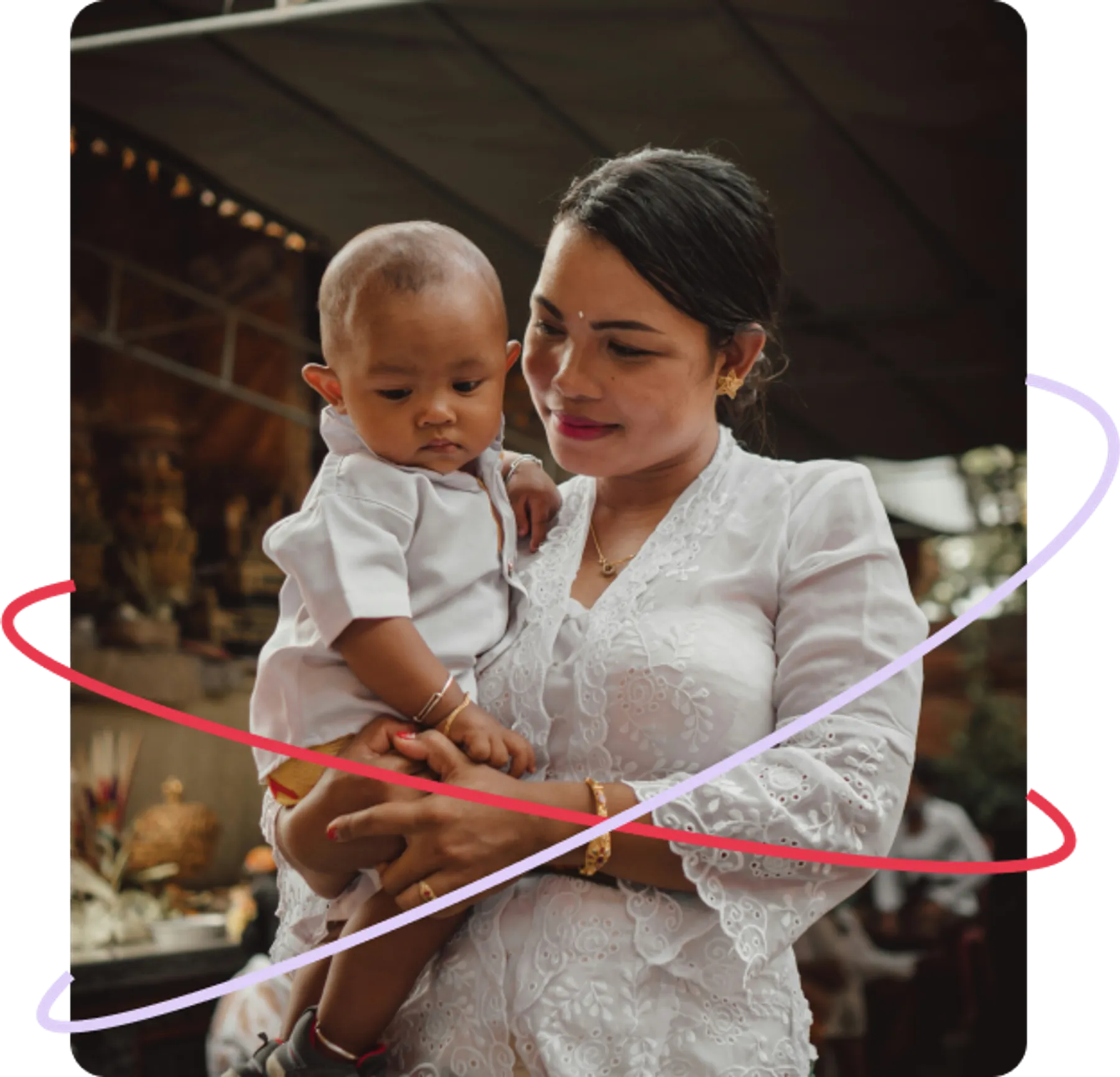 A mother dressed in a white embroidered top gently holds her young child in an indoor cultural setting, both looking down calmly.