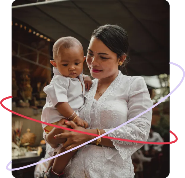 A mother dressed in a white embroidered top gently holds her young child in an indoor cultural setting, both looking down calmly.