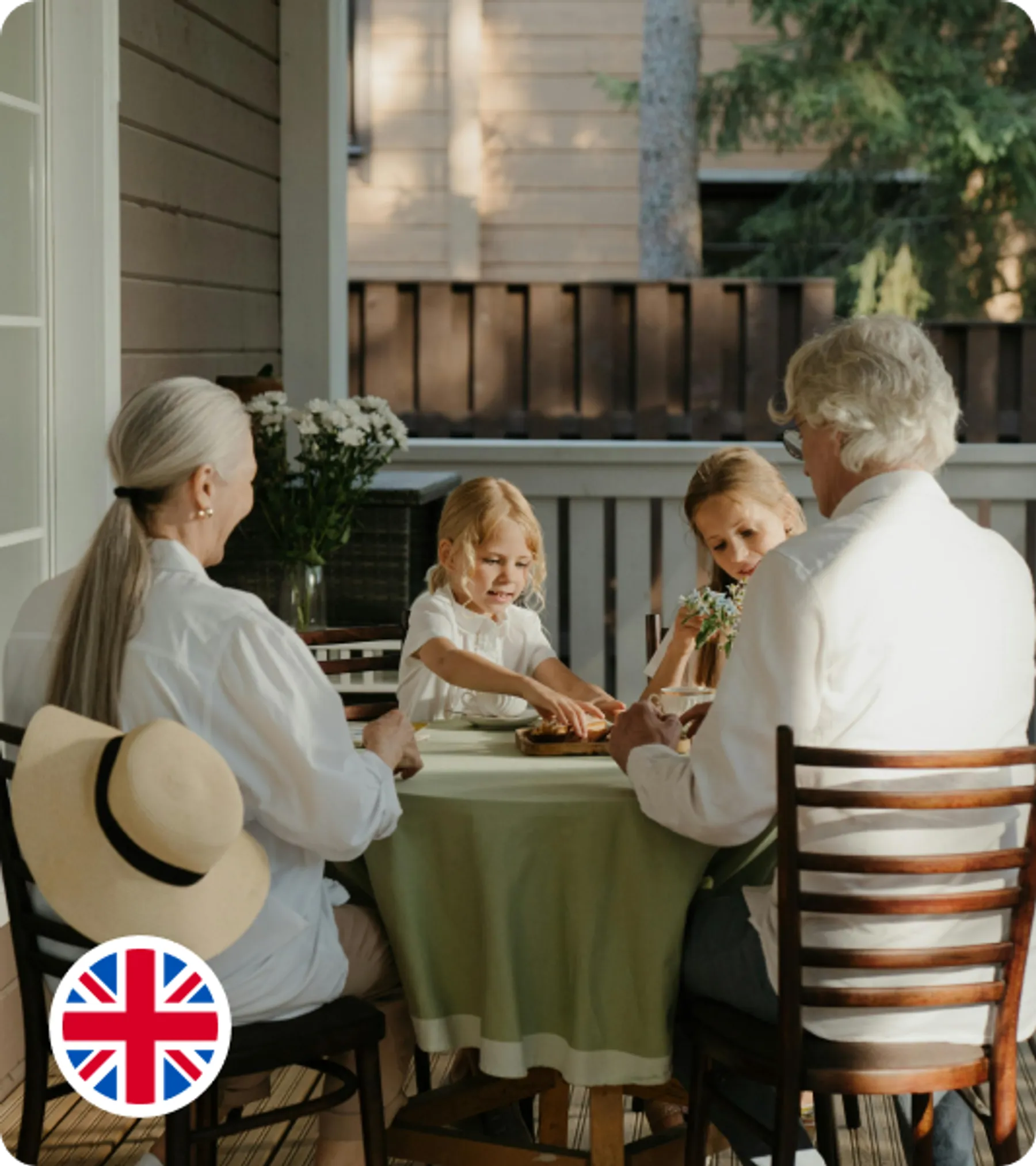 An elderly couple, two children, and a woman sit around a table on a porch, enjoying a meal together in soft daylight.