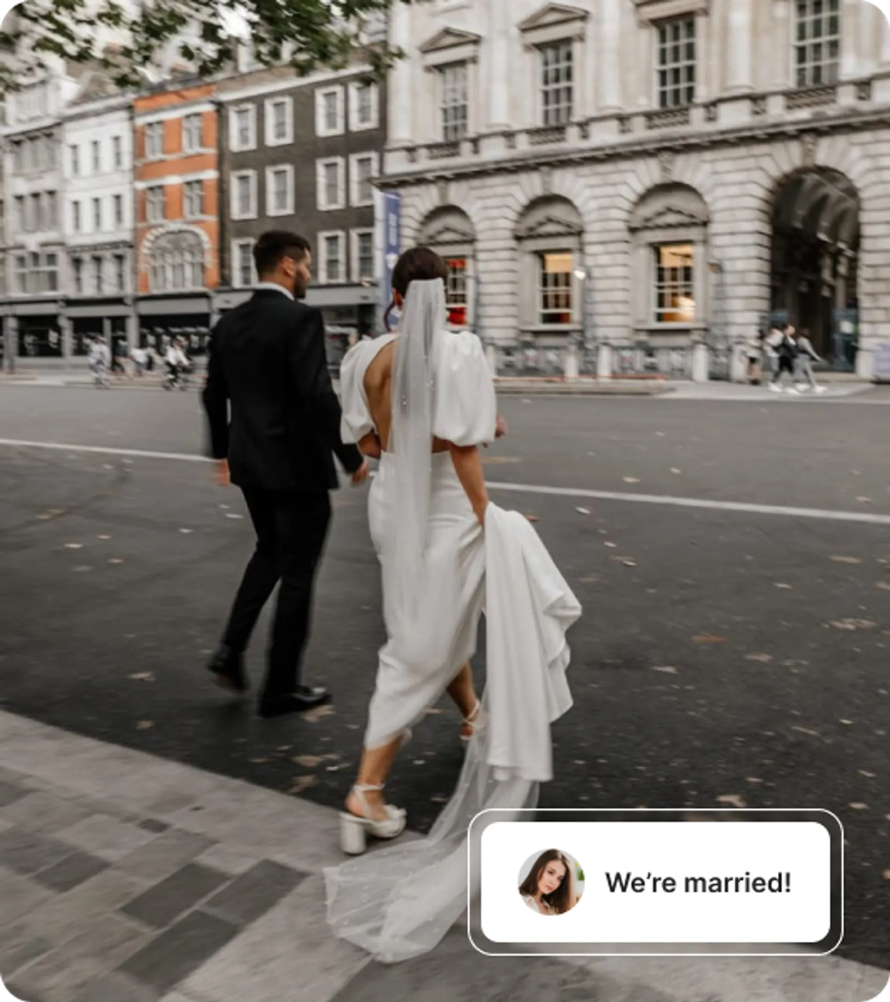 A bride in a flowing white dress and a groom in a dark suit walk hand-in-hand across a city street, celebrating their marriage.