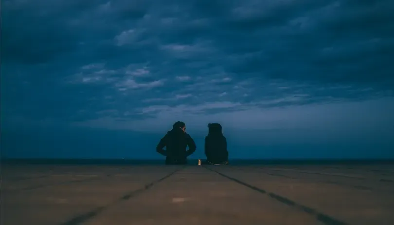 Distant couple sitting apart near the ocean under a moody sky, symbolizing emotional separation
