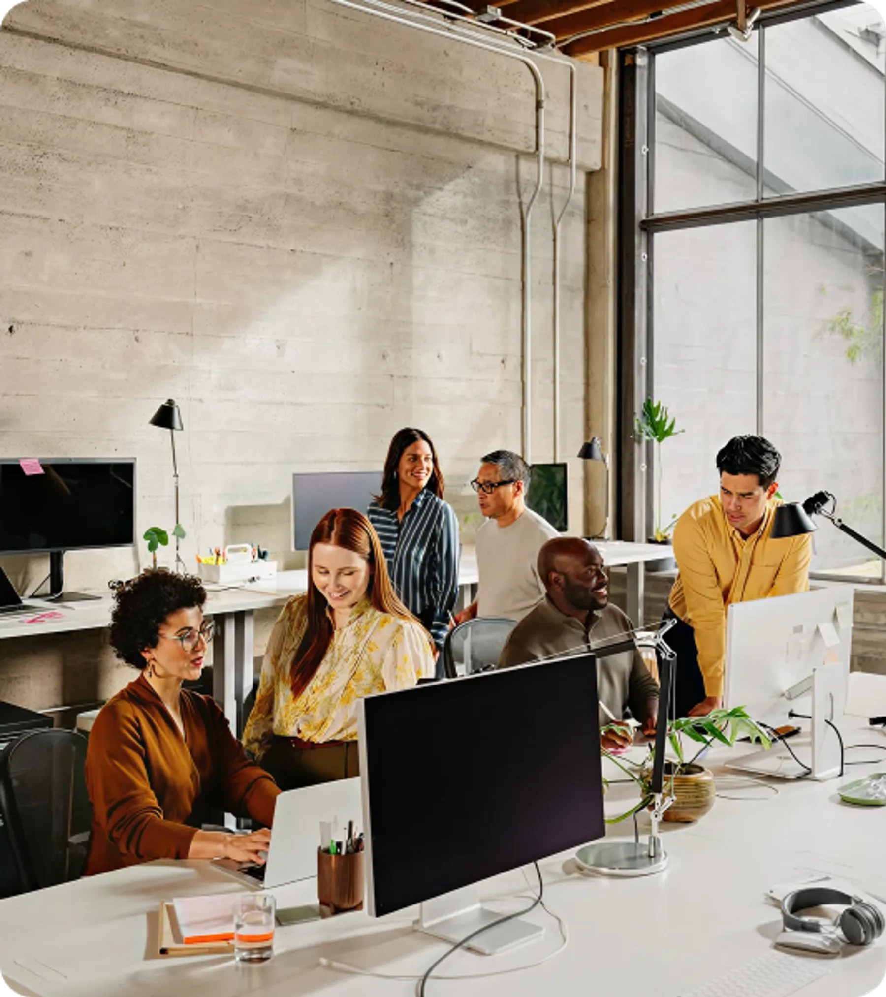 A group of professionals engaged in teamwork and conversation at a bright, open-plan tech startup office with large windows and computers.