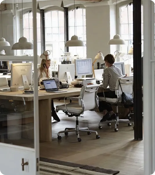 Several professionals working at computers in a bright, open-plan office with large windows and modern furnishings.