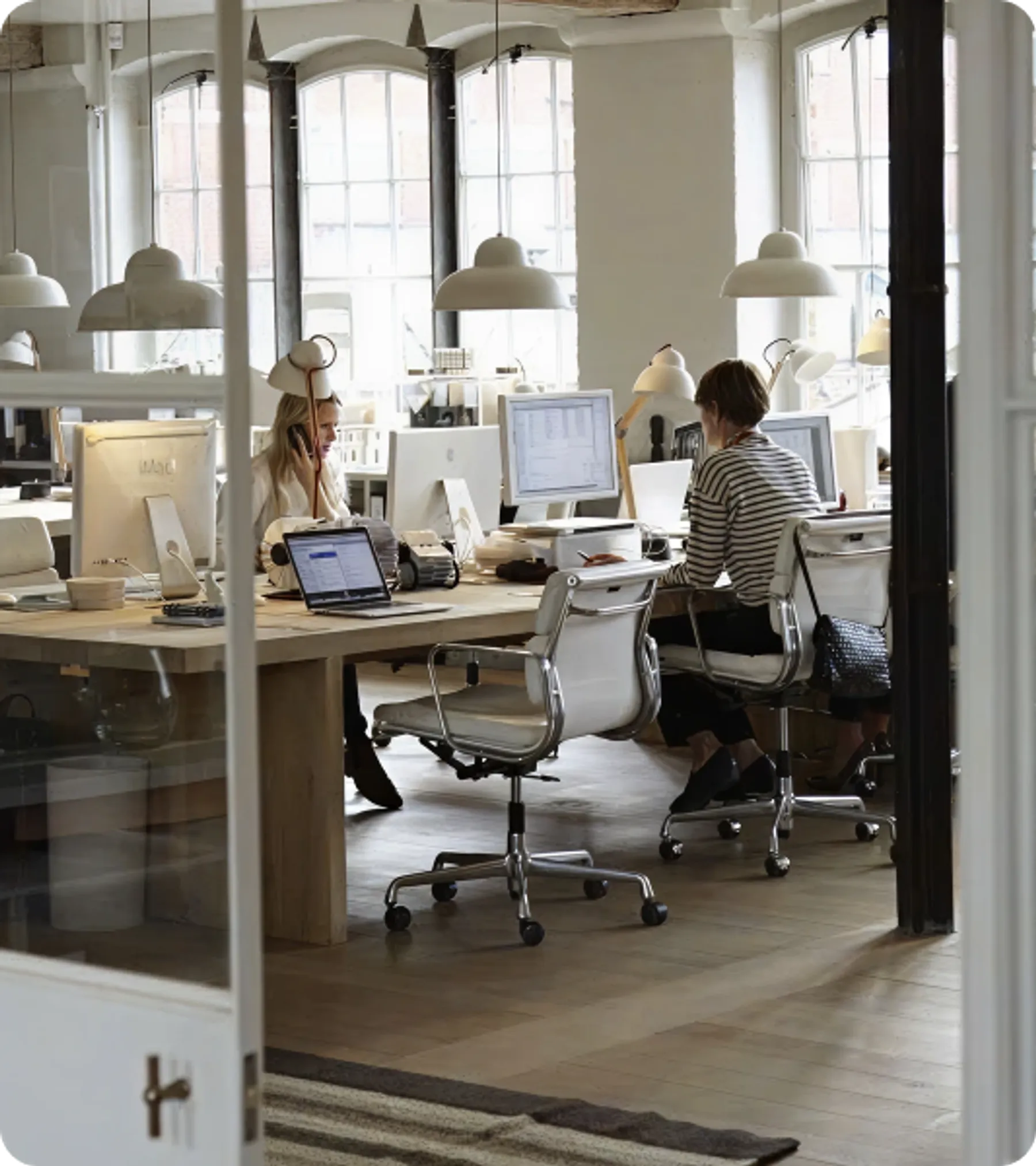 Several professionals working at computers in a bright, open-plan office with large windows and modern furnishings.