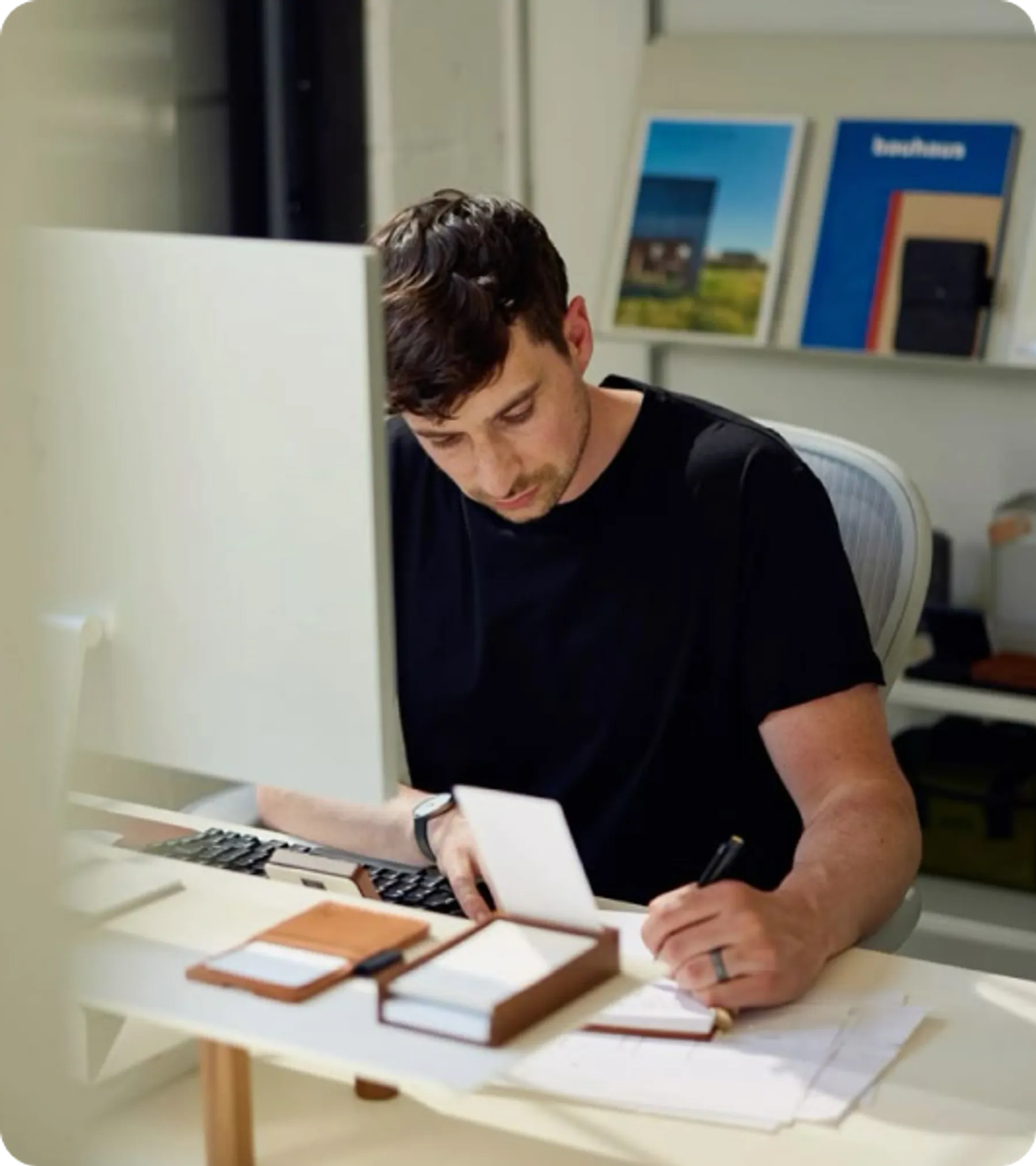 A man in a black shirt concentrating on paperwork and a desktop computer task in a modern office workspace.