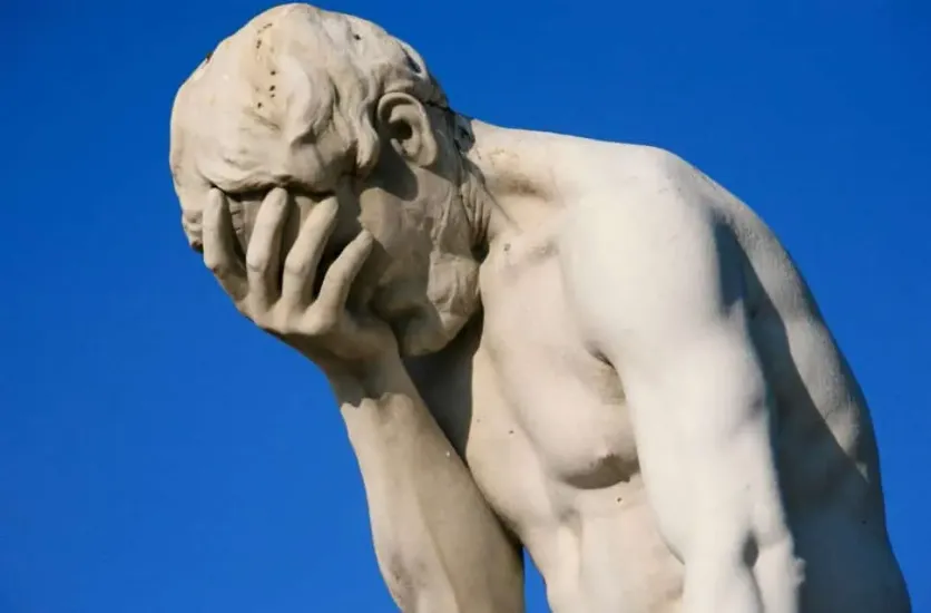 Statue of a man holding his face in his hand, expressing frustration or disappointment against a clear blue sky.