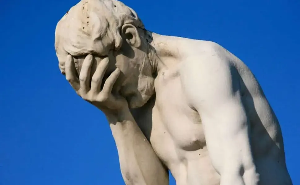 Statue of a man holding his face in his hand, expressing frustration or disappointment against a clear blue sky.