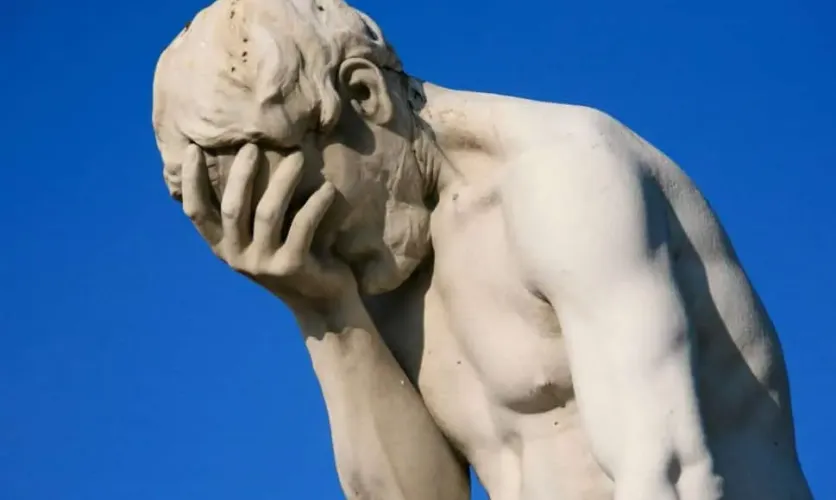 Statue of a man holding his face in his hand, expressing frustration or disappointment against a clear blue sky.