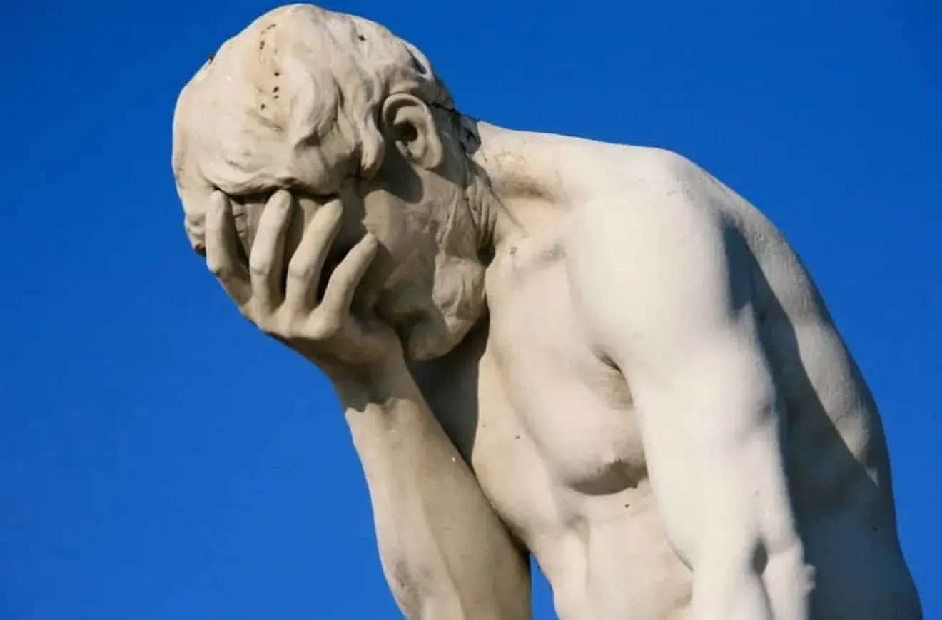 Statue of a man holding his face in his hand, expressing frustration or disappointment against a clear blue sky.