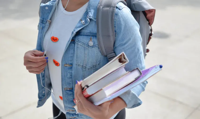 Student wearing a denim jacket and holding books with a backpack outdoors