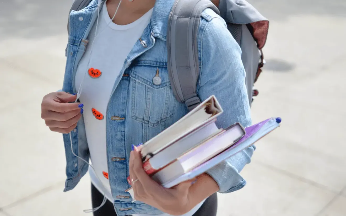 Student wearing a denim jacket and holding books with a backpack outdoors