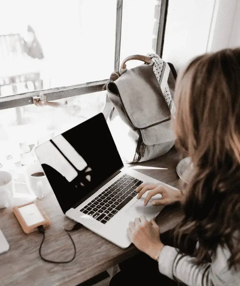 Woman working on a laptop by a window with a backpack beside her, representing digital nomad or remote work lifestyle