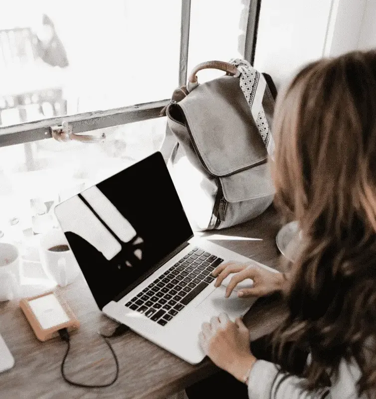 Woman working on a laptop by a window with a backpack beside her, representing digital nomad or remote work lifestyle