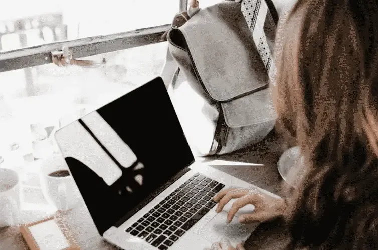 Woman working on a laptop by a window with a backpack beside her, representing digital nomad or remote work lifestyle