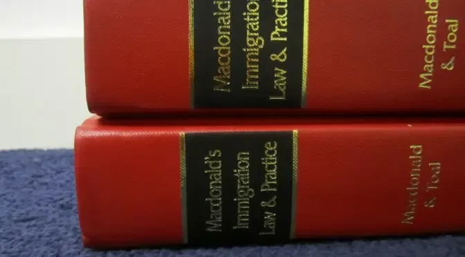 Two red-bound volumes of "Macdonald's Immigration Law & Practice" stacked on a dark surface, representing UK immigration legal reference