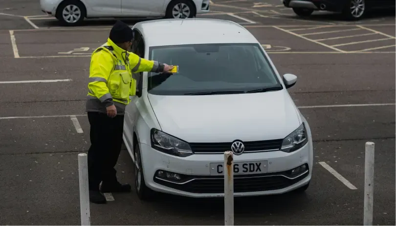 Traffic officer placing a parking penalty notice on a white car in a UK car park