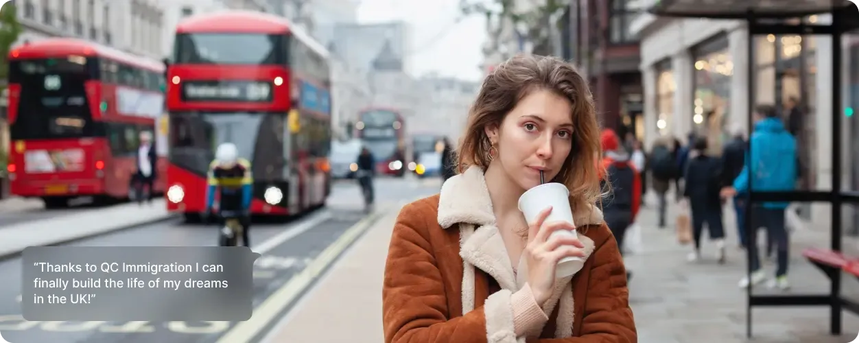 A woman in a brown shearling coat holding a coffee cup on a busy London street with red double-decker buses and pedestrians in the background.