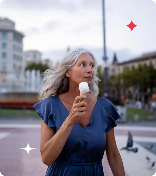 Woman with silver hair holding an ice cream cone while walking through a city square at dusk.